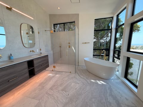 Modern luxury bathroom with freestanding white soaking tub, walk-in shower, dark wood vanity, and large windows overlooking trees