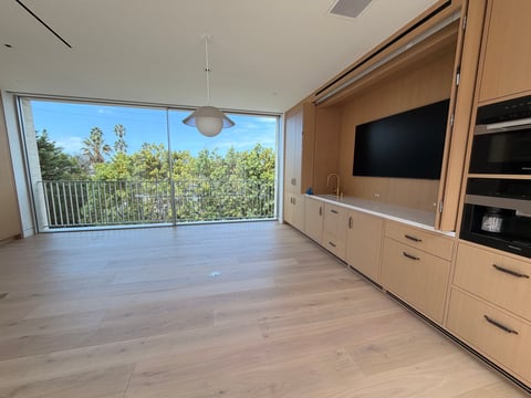 Modern apartment living room with light wood flooring, wall-mounted TV, built-in cabinetry, and floor-to-ceiling windows overlooking trees