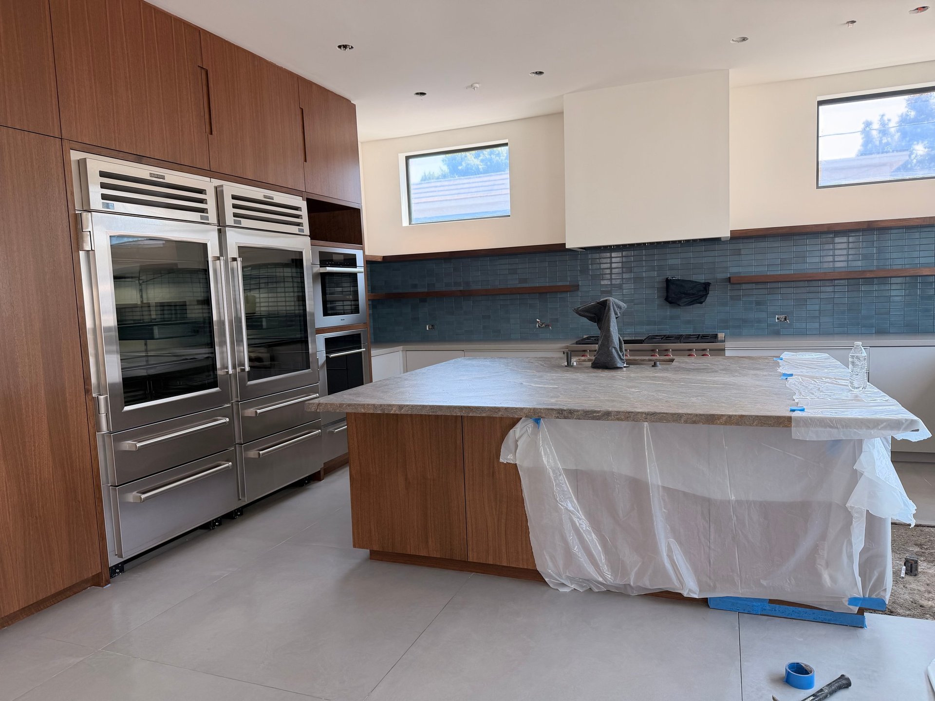 Modern kitchen under renovation with stainless steel appliances, wooden cabinetry, blue tile backsplash, and island counter covered in protective plastic sheeting