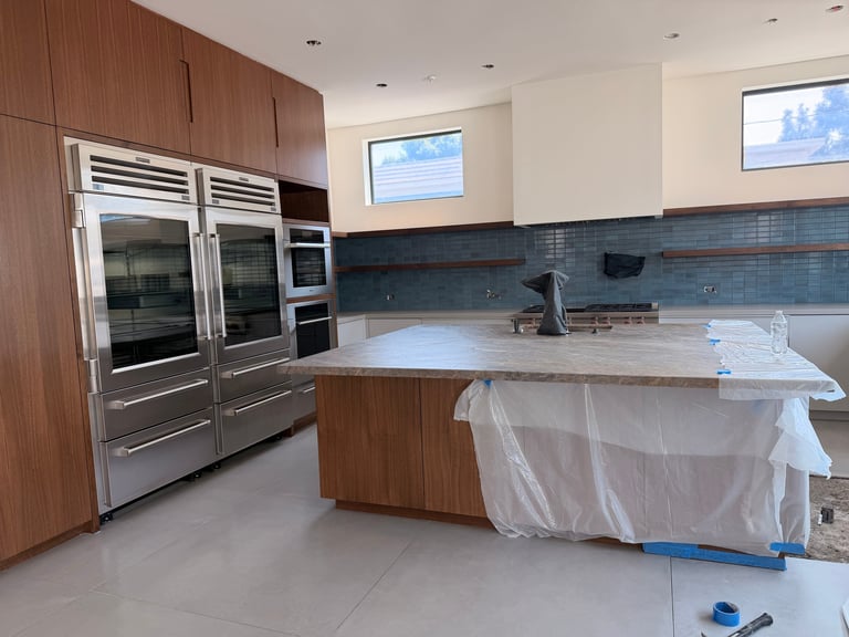 Modern kitchen under renovation with stainless steel appliances, wooden cabinetry, blue tile backsplash, and island counter covered in protective plastic sheeting
