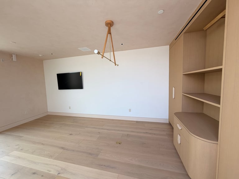 Empty room with wooden flooring, white walls, wall-mounted TV, wooden pendant light fixture, and built-in corner shelving unit