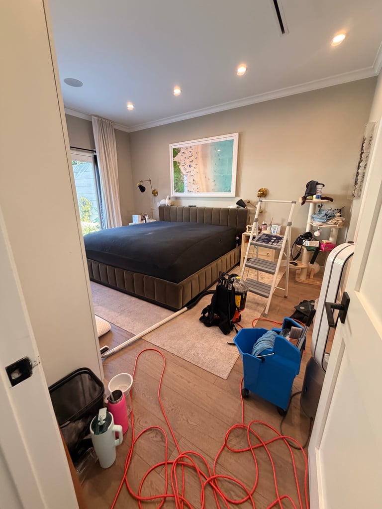 Modern bedroom with dark platform bed, sandy beige walls, window with beach artwork, red extension cords on wooden floor, and storage furniture along right wall.