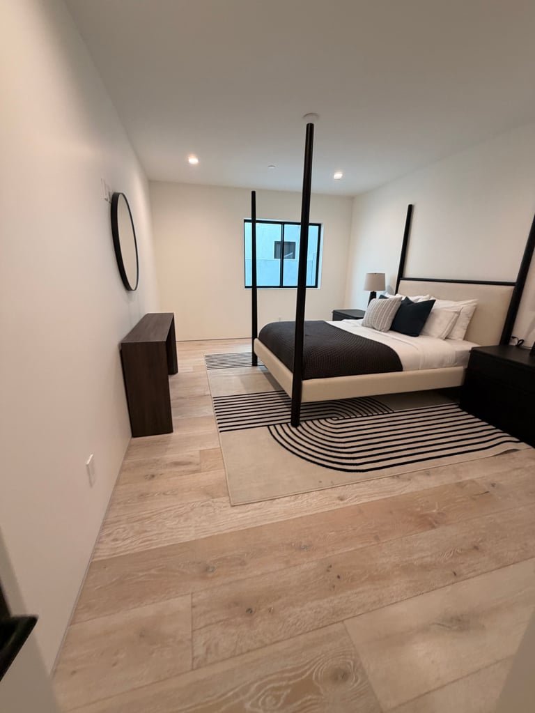 Modern bedroom with black four-poster bed, light wood flooring, geometric striped rug, mounted TV, and contemporary furnishings