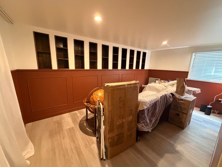 Bedroom interior with burgundy walls, wooden storage cabinets above, a bed with plastic covering, wooden furniture, and hardwood flooring.