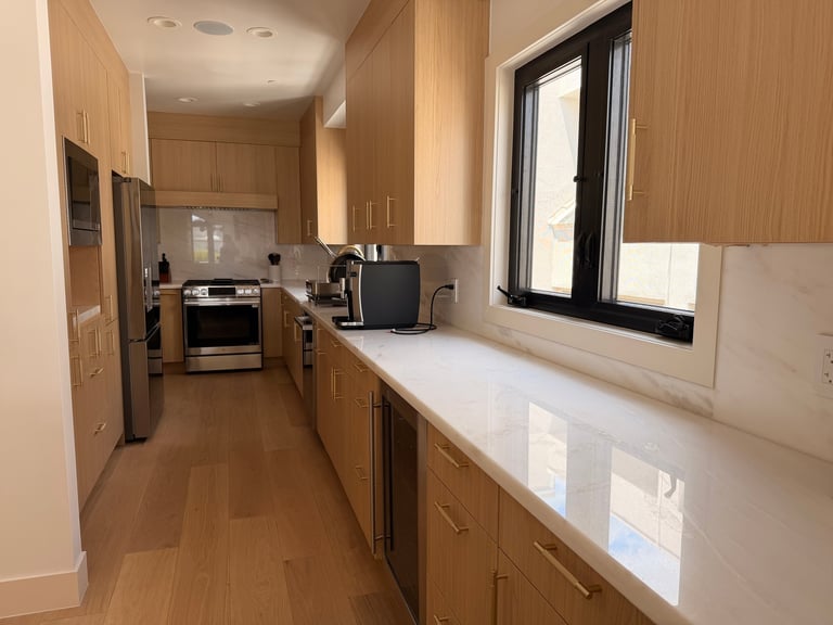 Modern galley kitchen with wooden cabinets, white countertop, stainless steel appliances, and a black-framed window