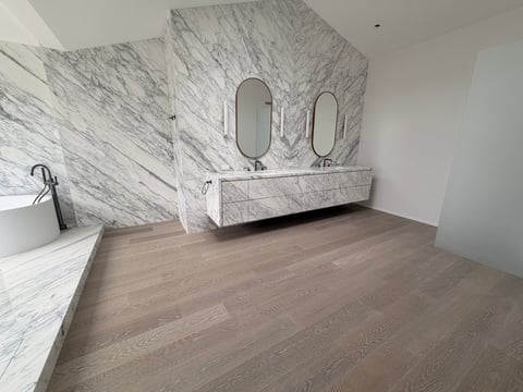 Modern bathroom with marble walls, double sink vanity, two oval mirrors, and wooden floor