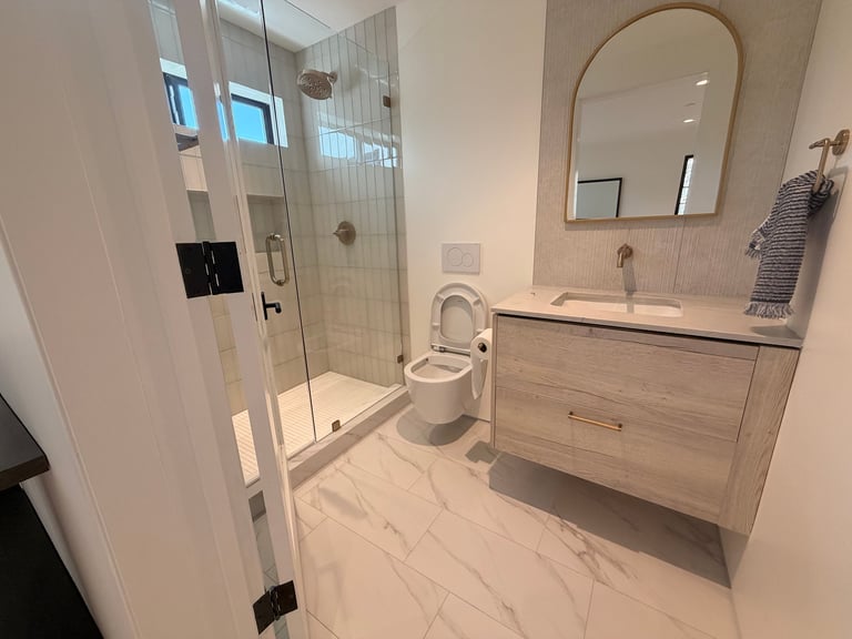 Modern bathroom with walk-in shower, white vanity, toilet, arched mirror, and marble tile flooring