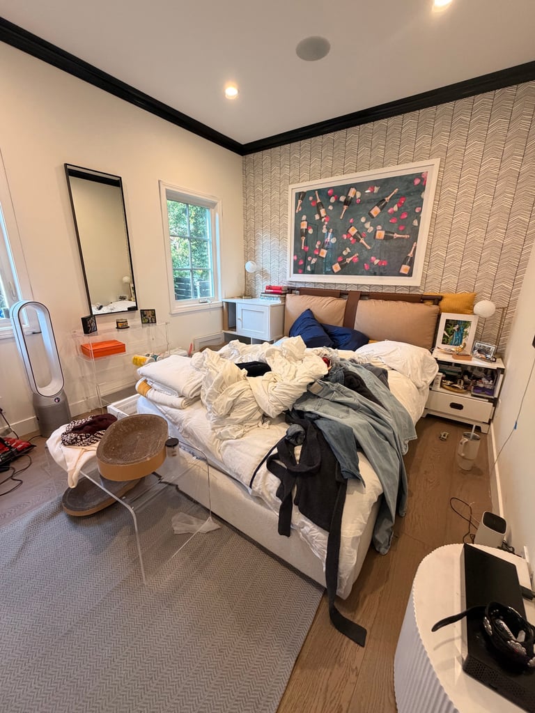 Messy bedroom with unmade bed, piles of clothes, white walls with textured wallpaper, framed artwork, and wooden flooring