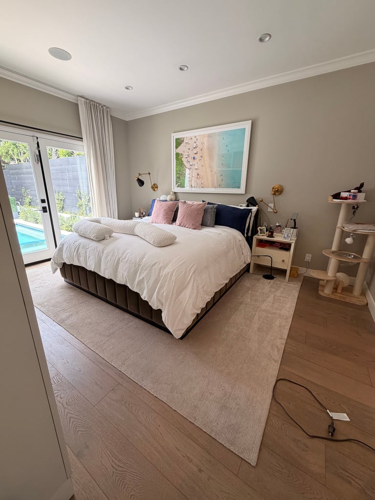 Modern bedroom with white bedding, pink and blue artwork, recessed ceiling lights, glass doors to pool area, and neutral taupe walls